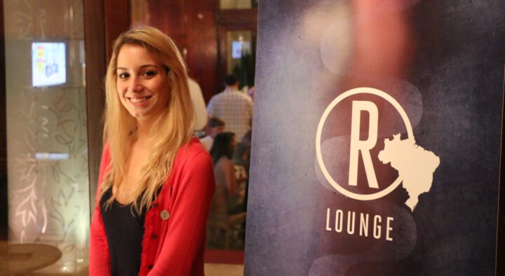Smiling woman in red stands next to an 'R Lounge' sign, emphasizing customer service in a relaxed setting at Roadtrips; discover and connect in welcoming environments.