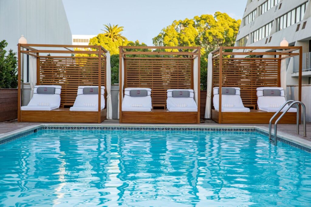 An outdoor pool at Hotel Dena in Pasadena, featuring clear blue water with sun loungers under wooden canopies, reflects a perfect relaxation spot for guests visiting during the Rose Bowl event.