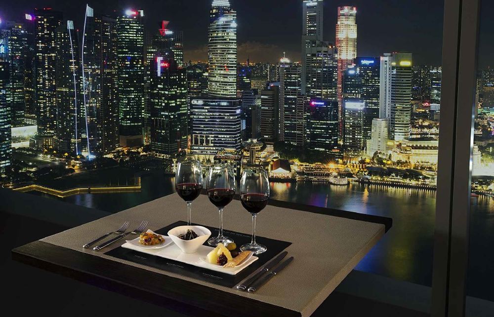 Dining setup with a sweeping night view of Singapore's skyline, showcasing glasses of wine and a meal, ideal for guests staying at nearby luxury hotels during the Singapore Grand Prix.