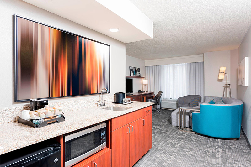 Spacious suite in the Courtyard by Marriott Louisville Downtown, featuring a modern kitchenette, large artistic wall art above the sink, and a cozy sitting area, ideal for Kentucky Derby accommodations.