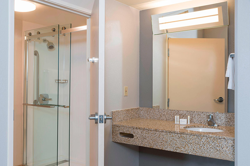 Modern bathroom at the Courtyard by Marriott Louisville Downtown, featuring a glass shower, large mirror, and granite vanity, ideal for visitors attending the Kentucky Derby.