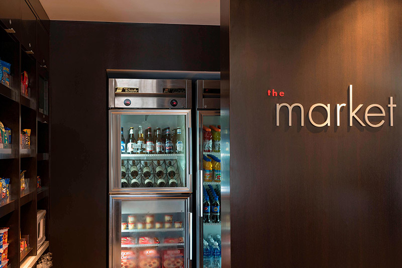 Inside the Courtyard by Marriott in downtown Louisville, a beverage cooler is fully stocked next to shelving with snacks, located beside a wall labeled 'the market,' ideal for Kentucky Derby visitors.