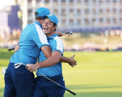 Two golfers in blue team uniforms joyfully hug on a golf course, capturing the camaraderie at events like the Ryder Cup, ideal for those exploring golf travel packages in Long Island, New York.