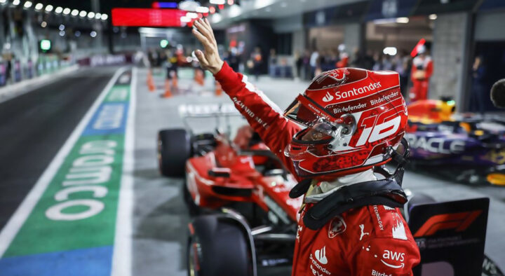 A Formula 1 driver in a red racing suit waves to the crowd during the Las Vegas Grand Prix, with a sleek race car and pit lane in the background.
