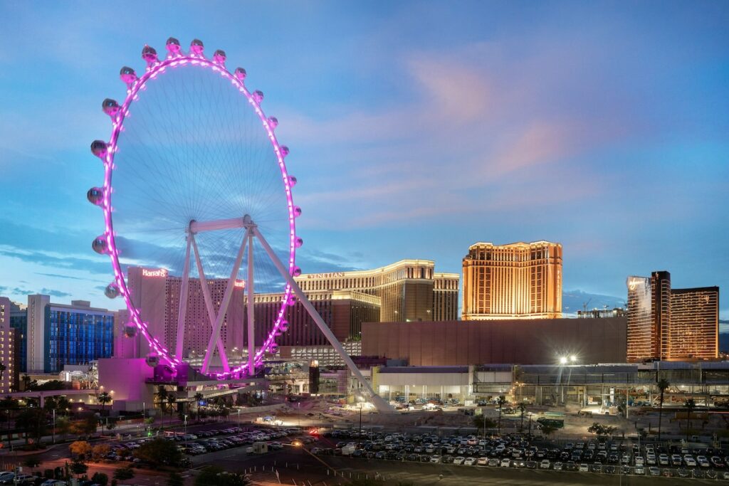 Twilight view of the Las Vegas skyline featuring the illuminated High Roller Ferris wheel, nearby hotels, and a bustling parking area, ideal for visitors of the Las Vegas Grand Prix.