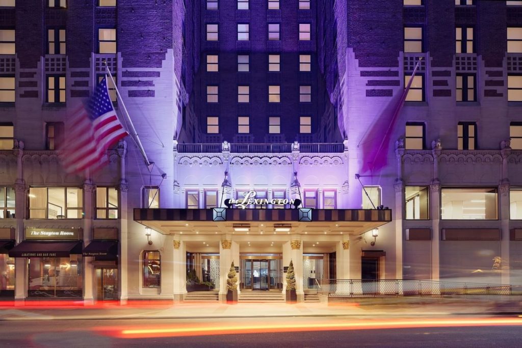 Evening view of the Lexington Hotel entrance illuminated by purple lights, featuring an American flag and bustling street scene, relevant for those interested in US Open Tennis Packages.