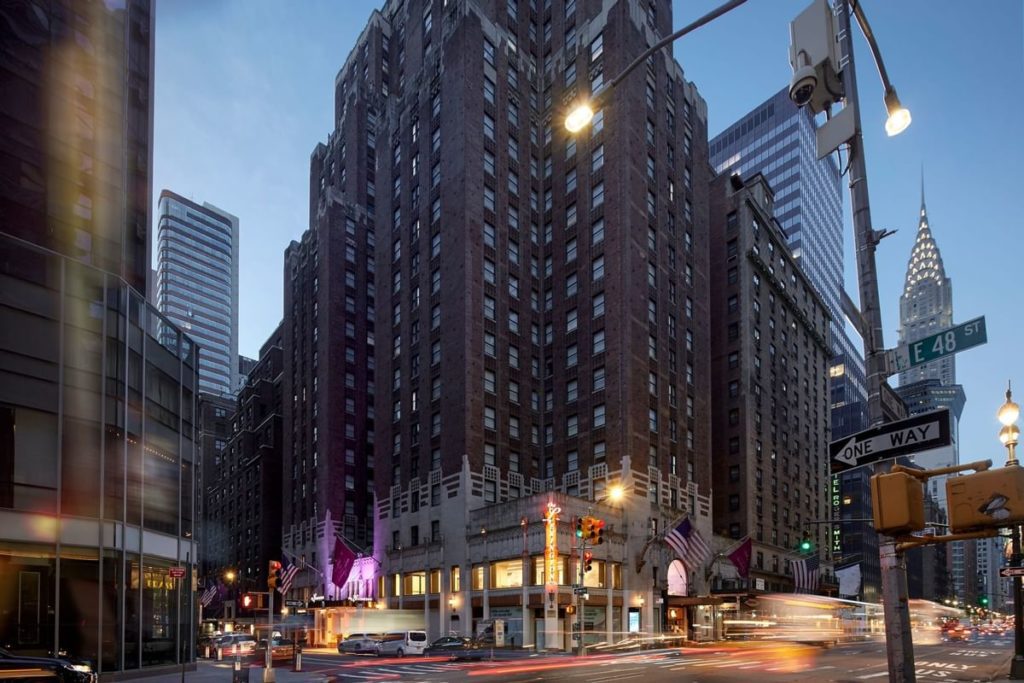 View of the Lexington Hotel in New York at dusk, showcasing its towering structure and vibrant street scene with nearby Chrysler Building visible, ideal as US Open Tennis accommodations.