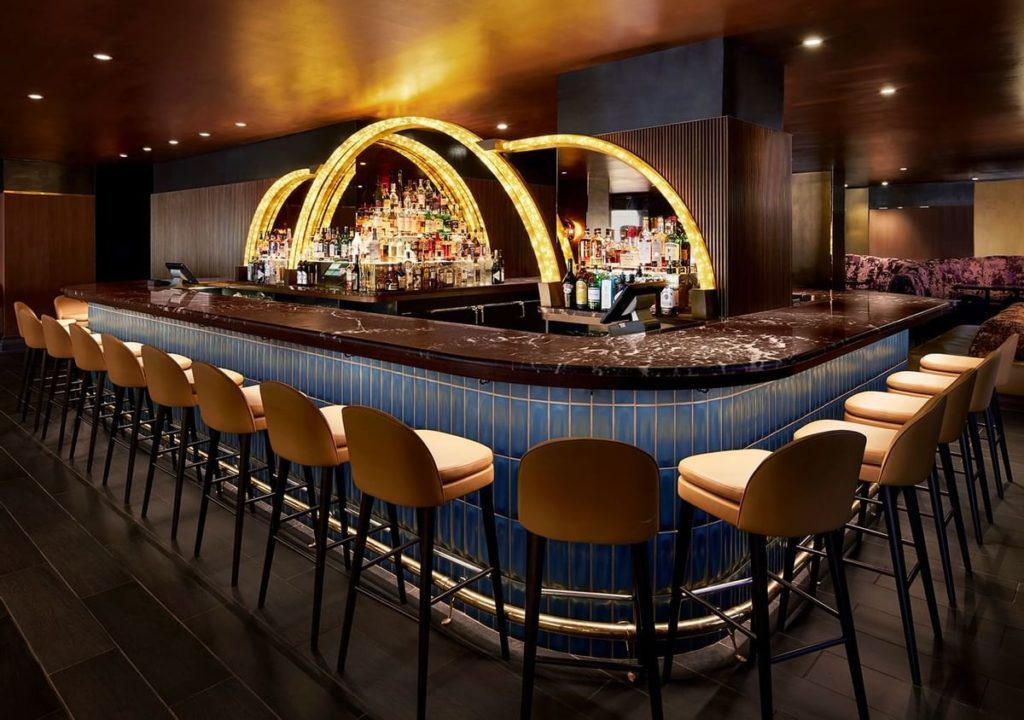 Luxurious bar at The Lexington Hotel, Autograph Collection in New York, featuring a curved counter with stools, golden arches, and a well-stocked back shelf, ideal for US Open Tennis fans.