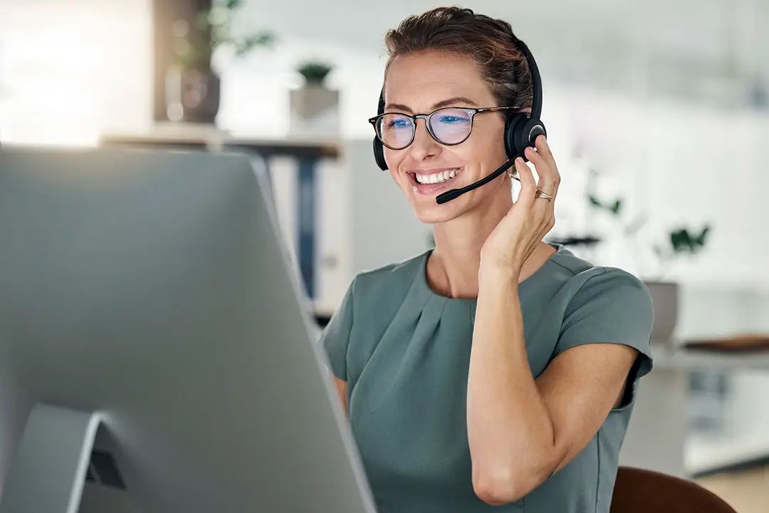 A customer service representative wearing a headset smiles while working at her computer in an office, associated with planning World Cup travel packages and event hosting services.