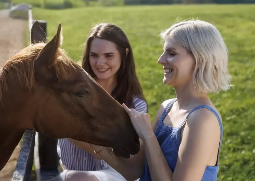 Two women, one petting a horse, enjoy a sunny day in a green pasture, perfect for those seeking premium Kentucky Derby packages that include travel and hotel accommodations.