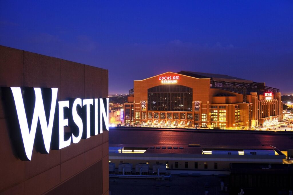 Night view showing the lit-up Westin hotel sign with the illuminated Lucas Oil Stadium in the background, suggesting a proximity to major Indianapolis attractions, ideal for visitors.