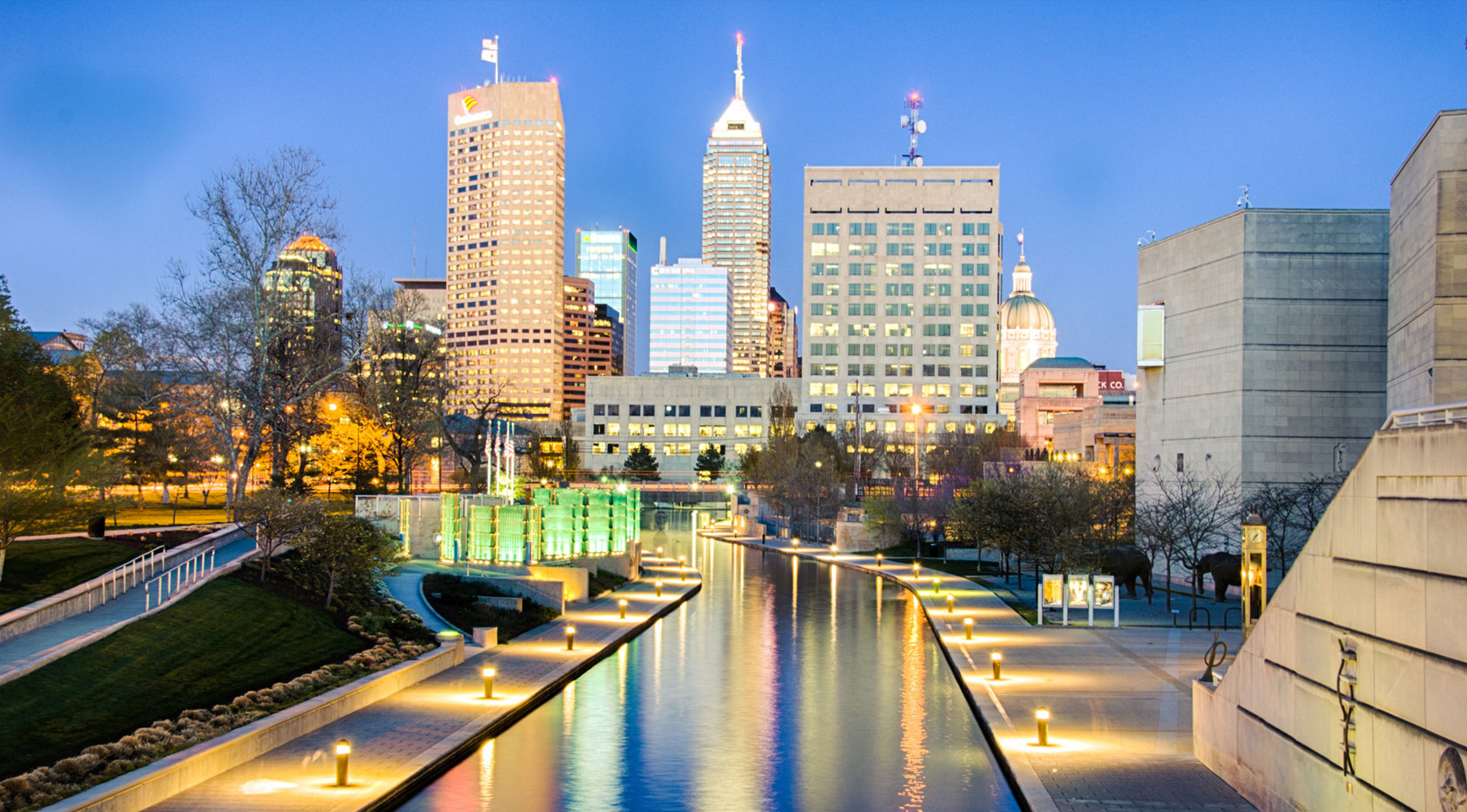 Evening view of downtown Indianapolis showcasing its illuminated skyline and the canal, a popular spot for tourists and locals alike, near the Indianapolis 500 area.