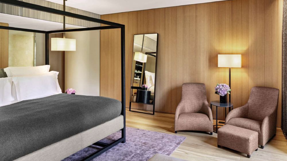 Interior view of a luxurious hotel room at Bvlgari Hotel Milano featuring a neatly made bed with a black canopy frame, brown armchairs, soft lighting, and wooden wall paneling, near Via Privata Fratelli Gabba.