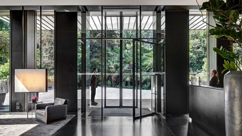 Modern lobby at Bvlgari Hotel Milano, showcasing sleek design with large glass doors offering a view of lush gardens, and a person entering through the doors. Ideal for guests visiting the Italian Grand Prix in Milan.