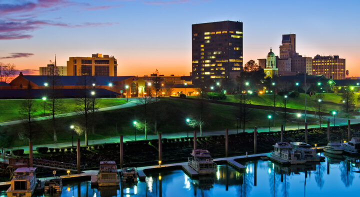 Twilight view of Augusta, Georgia, showing the serene riverfront lined with boats, illuminated walking paths, and modern city buildings, near the Augusta National Golf Club venue.