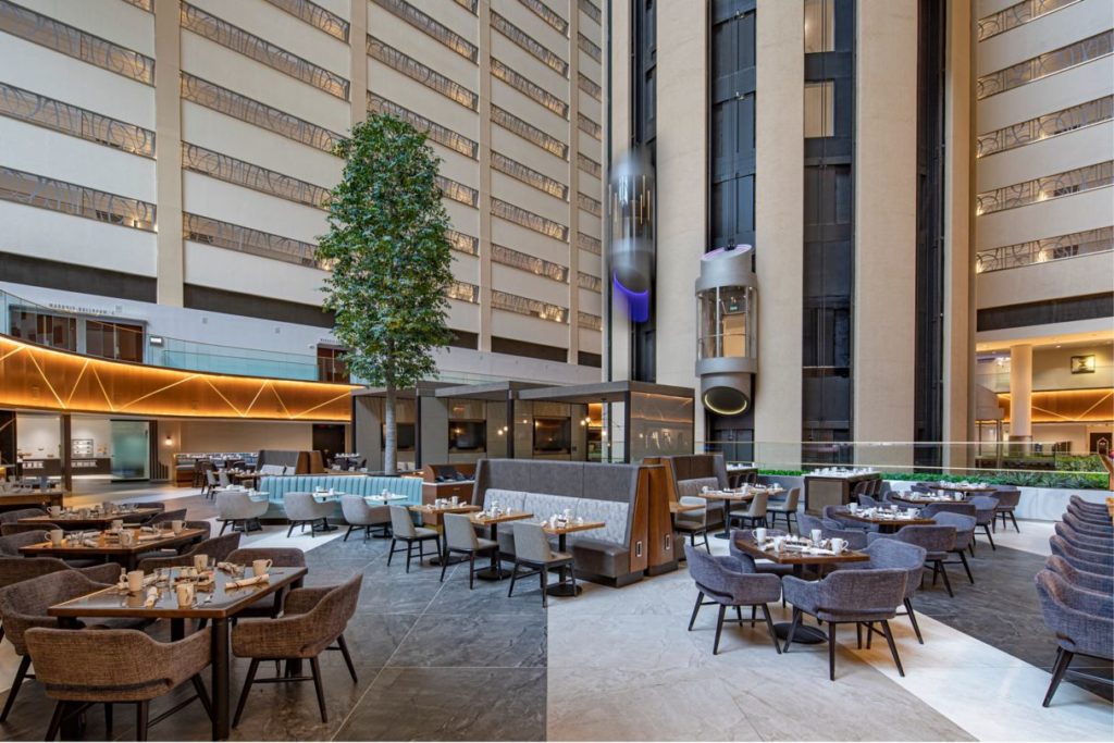 Luxurious atrium dining area inside the New York Marriott Marquis with multiple seating arrangements, set tables, and a towering central tree, ideal for US Open Tennis accommodation seekers.