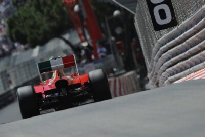 A race car speeds along the track at the iconic Monaco Grand Prix, with spectators and safety barriers lining the circuit.
