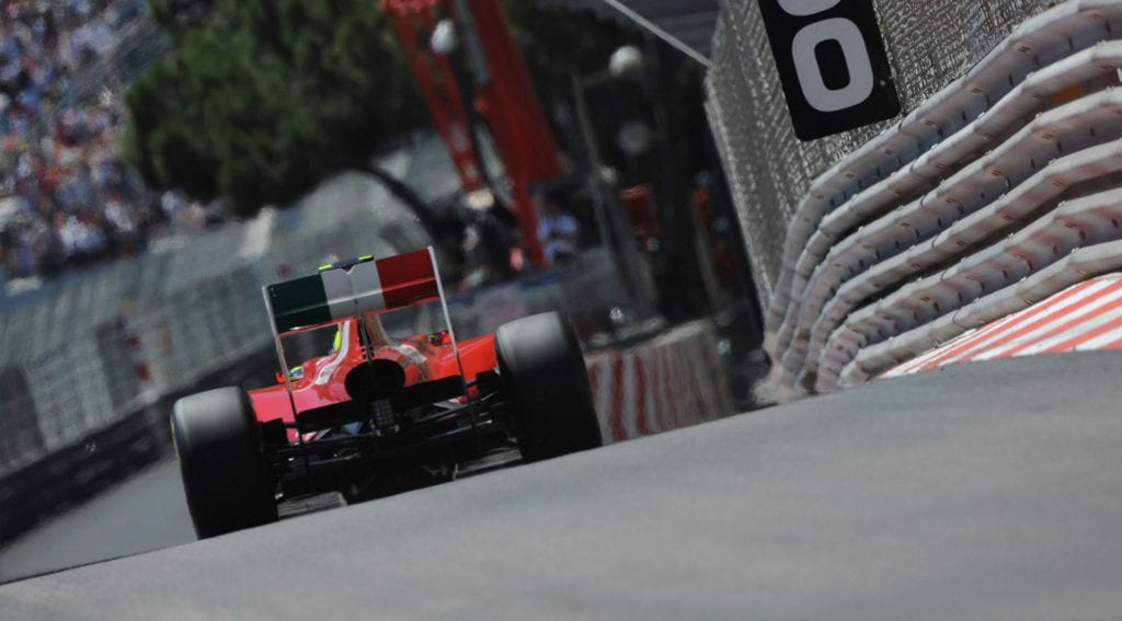 A race car speeds along the track at the iconic Monaco Grand Prix, with spectators and safety barriers lining the circuit.