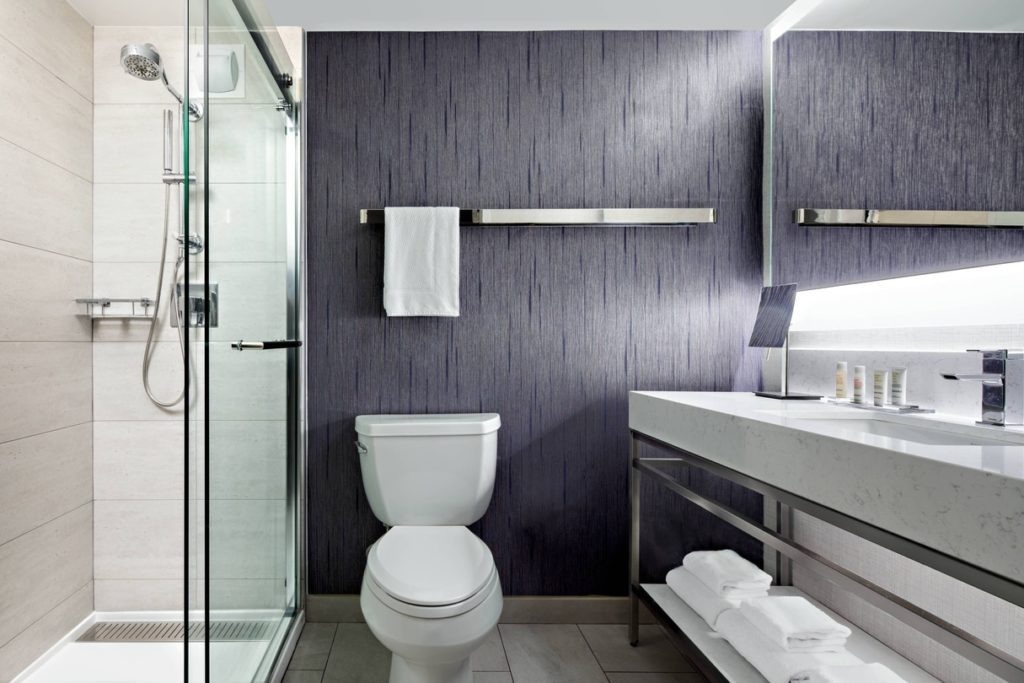 A modern hotel bathroom features a glass shower, gray wood-paneled walls, and a large vanity.