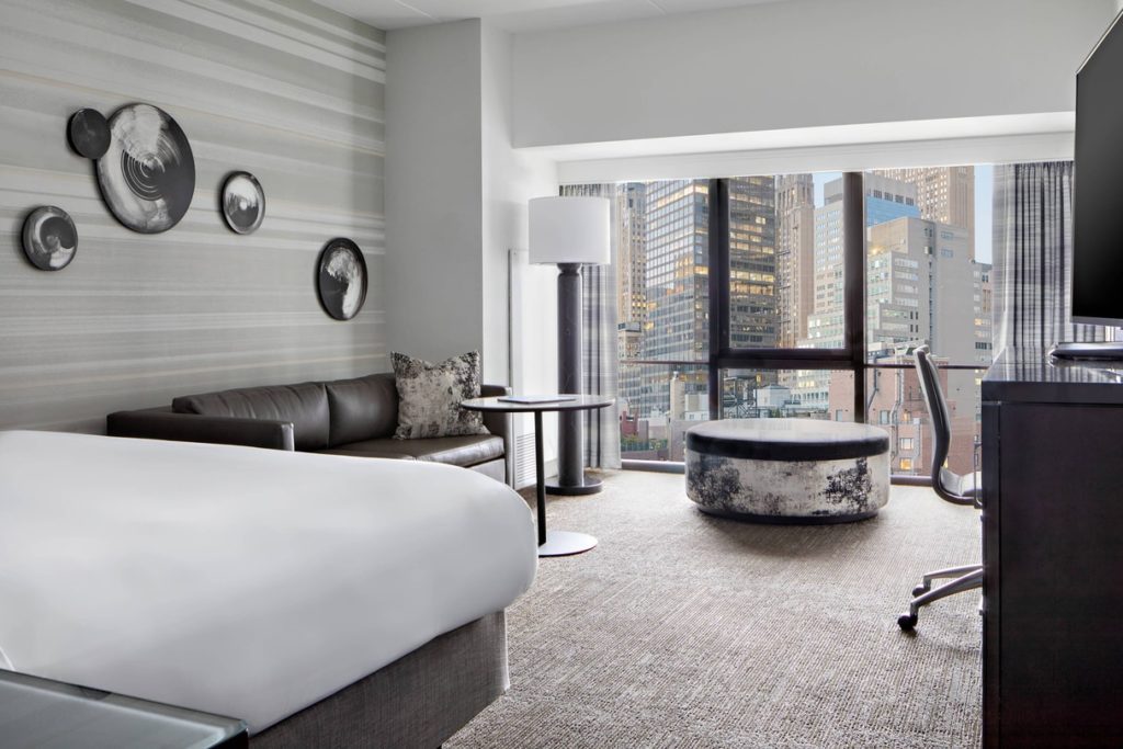A modern guest room featuring a large bed and comfortable seating, with a panoramic view of city skyscrapers, located at the New York Marriott Marquis, a recommended hotel for US Open Tennis spectators.