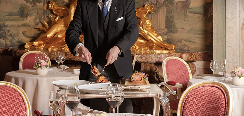 A waiter prepares food at an elegantly set table inside The Ritz London, ideal for guests attending Wimbledon events, showcasing the hotel's luxury dining experience.