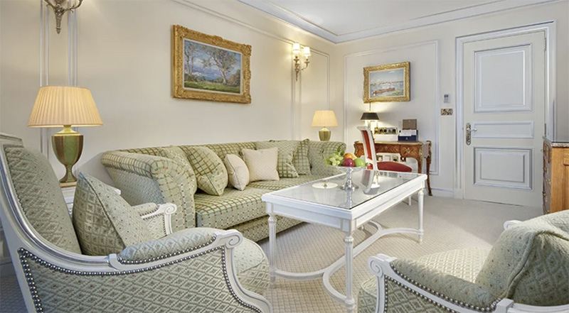 Elegant guest room at The Ritz London featuring plush green and white sofas, classical white furniture, ornate lamps, and framed landscape paintings, ideal for staying during the Wimbledon Championships.