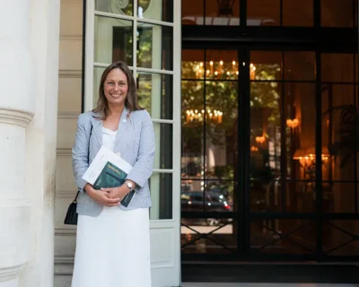 A woman stands smiling outside a building entrance, holding documents, representing personalized pre-trip assistance and high-quality sports travel experiences from Roadtrips' At-Your-Service Desk™.