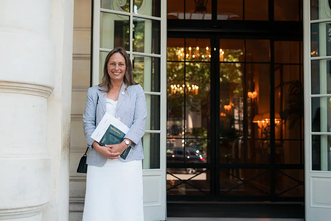 A woman stands smiling outside a building entrance, holding documents, representing personalized pre-trip assistance and high-quality sports travel experiences from Roadtrips' At-Your-Service Desk™.