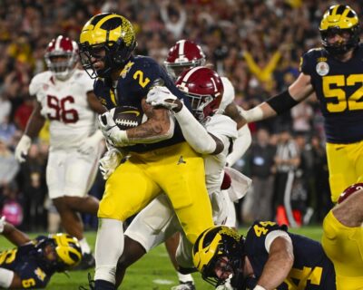 A football player from Michigan in a yellow uniform races forward with the ball while being tackled by an Alabama player in a high-tension game, capturing the dynamic essence of Rose Bowl action.