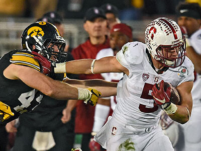 Two football players in a stadium are engaged in a play during a night game, promoting Rose Bowl travel packages that include the parade and luxury tours.