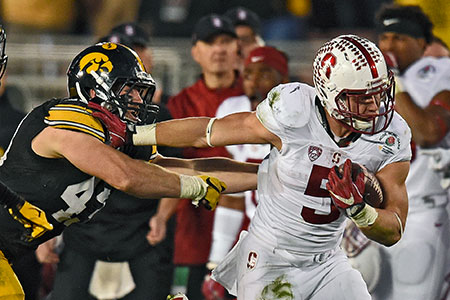 Two football players in a stadium are engaged in a play during a night game, promoting Rose Bowl travel packages that include the parade and luxury tours.