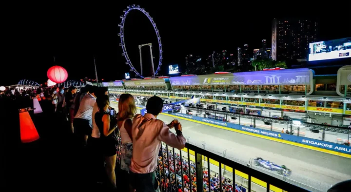 Spectators watch a night race at the Singapore Grand Prix, featuring illuminated tracks and the Singapore Flyer Ferris wheel in the background.