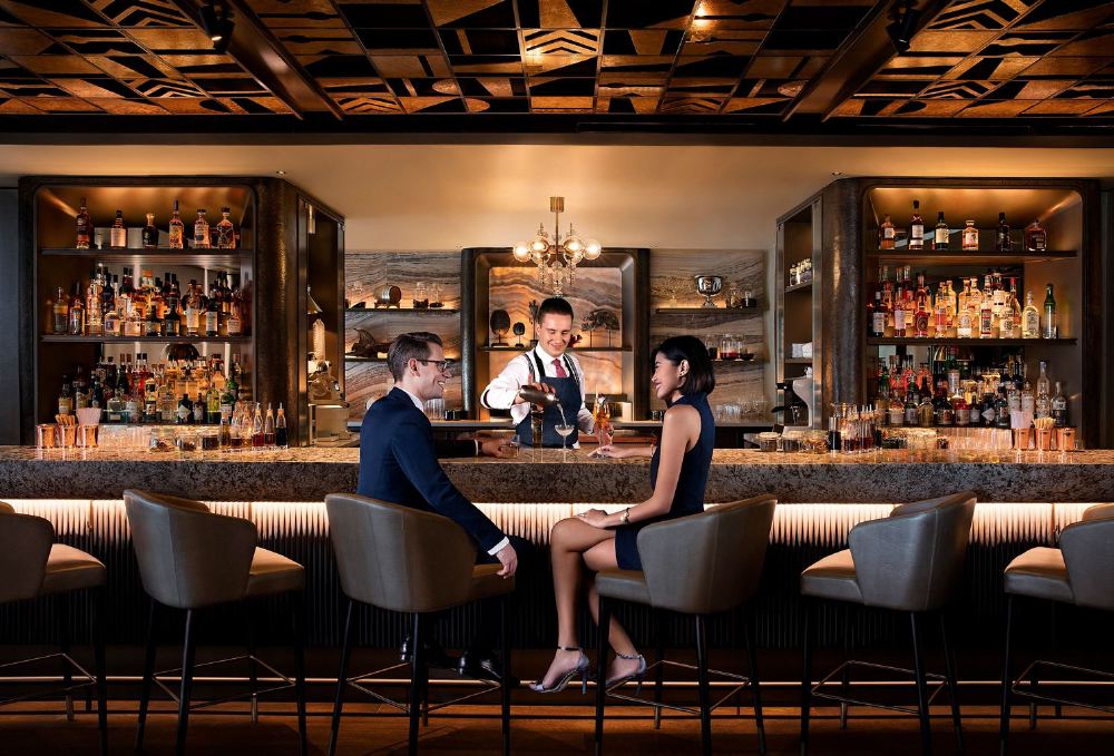 A couple enjoys drinks served by a bartender at MO BAR at Mandarin Oriental in Singapore, showcasing a sophisticated setting ideal for visitors attending the Singapore Grand Prix.