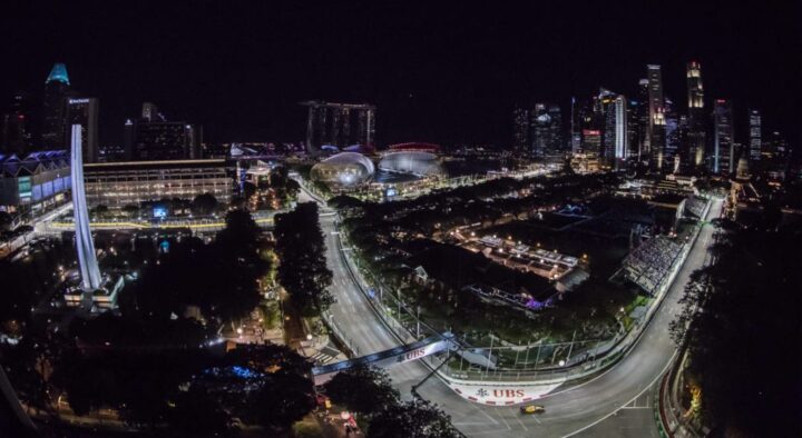 Nighttime aerial view of the Singapore Grand Prix circuit brightly lit with city skyscrapers in the background and a racecar speeding along the track.