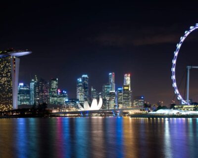 Nighttime view of Singapore's illuminated skyline featuring iconic structures like Marina Bay Sands and the Singapore Flyer, reflecting on the water.