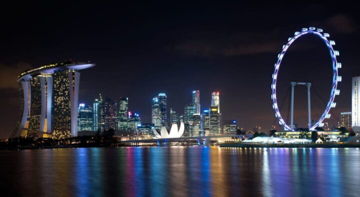 Nighttime view of Singapore's illuminated skyline featuring iconic structures like Marina Bay Sands and the Singapore Flyer, reflecting on the water.