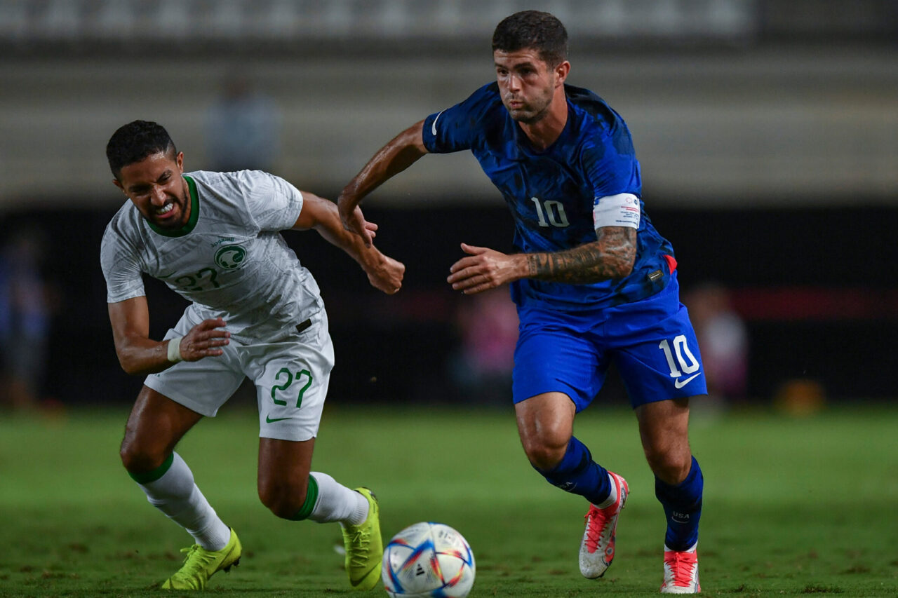Two soccer players compete for the ball during a match; one is clad in white, the other in blue, intensively focused on gaining possession in a stadium setting.