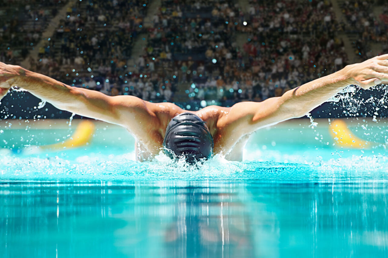 Male swimmer executing a butterfly stroke in a competition pool, surrounded by a crowd, capturing the intensity and skill of aquatic sports at the Summer Games.