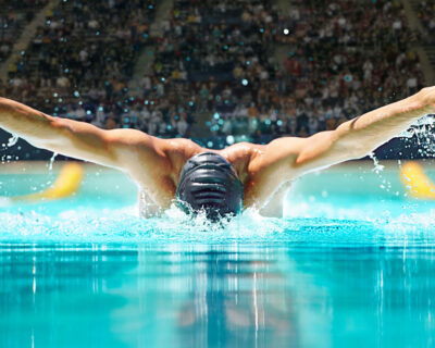 Male swimmer executing a butterfly stroke in a competition pool, surrounded by a crowd, capturing the intensity and skill of aquatic sports at the Summer Games.