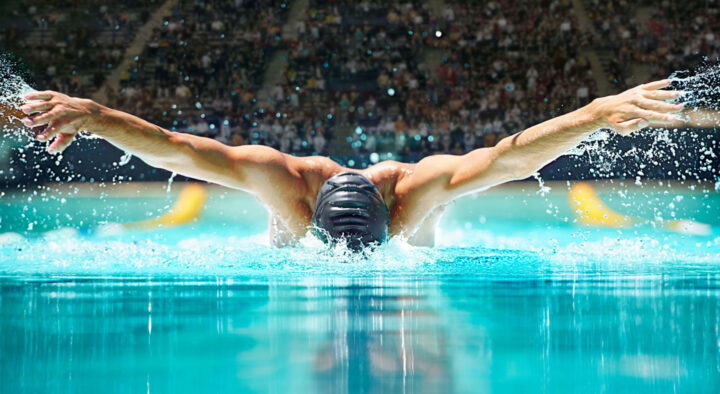 Male swimmer executing a butterfly stroke in a competition pool, surrounded by a crowd, capturing the intensity and skill of aquatic sports at the Summer Games.