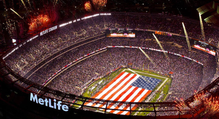 Interior view of a packed MetLife Stadium during an event, featuring a large American flag on the field and fireworks.
