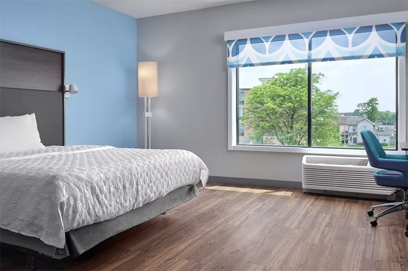 Modern hotel room at Tru by Hilton Indianapolis Downtown featuring a stylish gray bed, blue walls, and large window with view, ideal for guests visiting Indianapolis, Indiana for the Indianapolis 500.