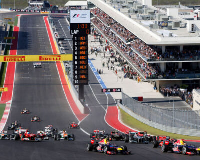 Race cars speed around a track at the US Grand Prix, with a large leaderboard displaying race standings and crowds filling stadium seats, highlighting the excitement of motorsport events.