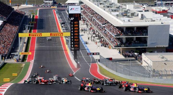 Race cars speed around a track at the US Grand Prix, with a large leaderboard displaying race standings and crowds filling stadium seats, highlighting the excitement of motorsport events.