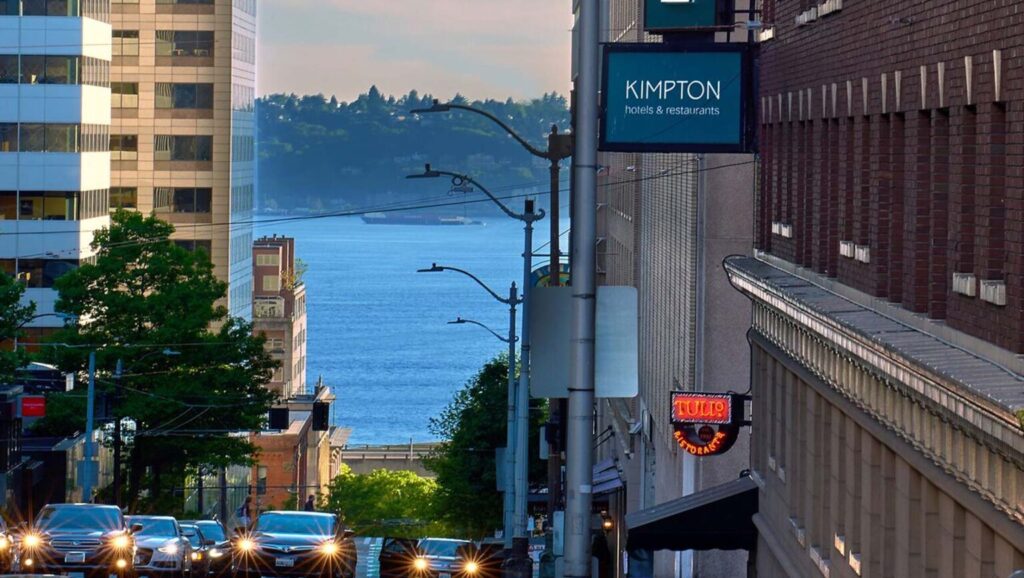 View of Puget Sound visible from a city street lined with buildings, where a Kimpton hotel sign is prominently displayed, in Seattle, Washington.