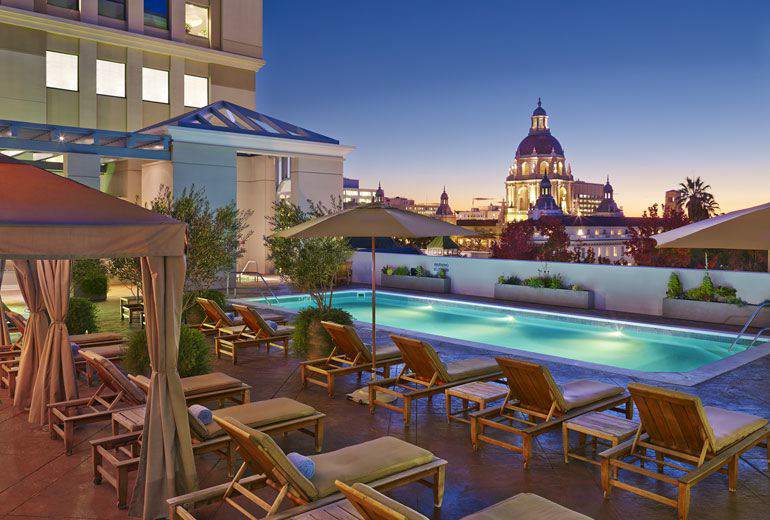 Luxury hotel pool area in Pasadena, California, with lounge chairs and illuminated pool, showcasing a view of a grand dome-topped building in the background at dusk.