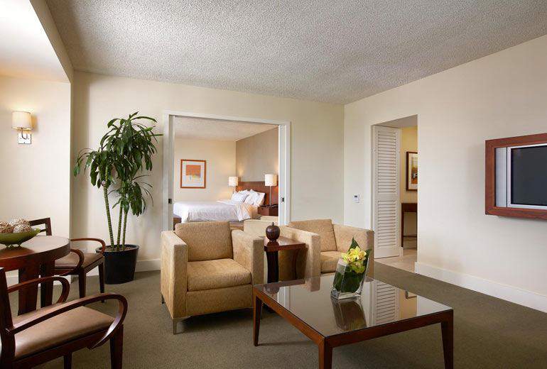 Spacious hotel suite at the Westin Pasadena featuring a living area with beige sofas, a glass coffee table, and a separate bedroom area, ideal for Rose Bowl visitors in Pasadena, California.