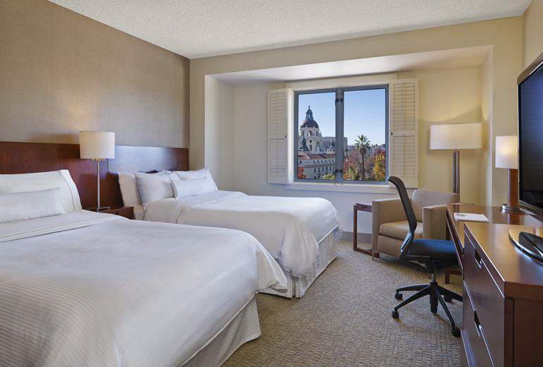 An elegant hotel room at Westin Pasadena featuring two queen-sized beds, a work desk, and a large window with a view of a historic building, ideal for visitors to the Rose Bowl in Pasadena.