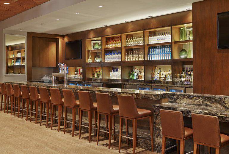 Elegant bar setup at Westin Pasadena, featuring a long marble counter with rows of brown leather stools, well-stocked shelves, and rich wooden décor, ideal for Rose Bowl visitors.