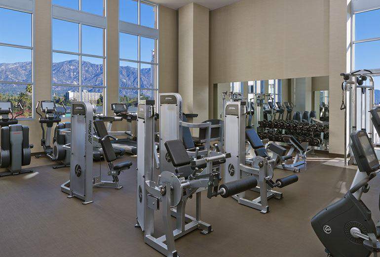 Modern fitness center equipped with state-of-the-art exercise machines, set against the backdrop of large windows showcasing mountain views, located at Westin Pasadena, a luxury hotel near the Rose Bowl in Pasadena, California.